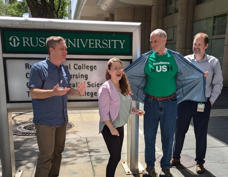 The TC Core Team (Paul Galchutt, Cate Beaulieu-Desjardins, George Fitchett, and Csaba Szilagyi) in front of a Rush University sign. George is wearing a Rush T-Shirt and the whoel team is smiling and laughing.